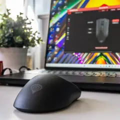 Czarna myszka komputerowa z logo Genesis na biurku, obok laptopa z kolorową tapetą i oprogramowaniem do konfiguracji. Wygląda jak sprzęt refurbished.