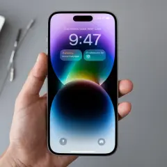 Dłoń trzyma iPhone 14 z kolorową tapetą i widoczną godziną 9:47. Ekran odświeżanie jest płynne.