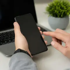 Dłoń trzyma smartfon, palec wskazujący dotyka ekranu, obok laptop. Może to być moment przed przywróceniem ustawień fabrycznych.