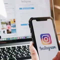 Dłoń trzyma smartfon z logo Instagrama. Obok laptop z otwartą stroną logowania, pokazującą jak wylogować się z Instagrama.