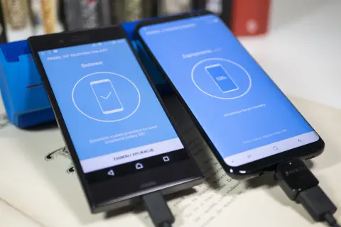 Dwa telefony połączone kablem USB. Jeden z nich pokazuje komunikat "Gotowe", drugi "100% Aktualizacja danych Kontakty...", co jest częścią procesu jak uruchomić nowy telefon Samsung.