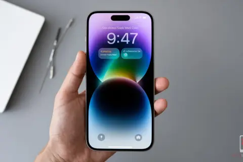 Dłoń trzyma iPhone 14 z kolorową tapetą i widoczną godziną 9:47. Ekran odświeżanie jest płynne.
