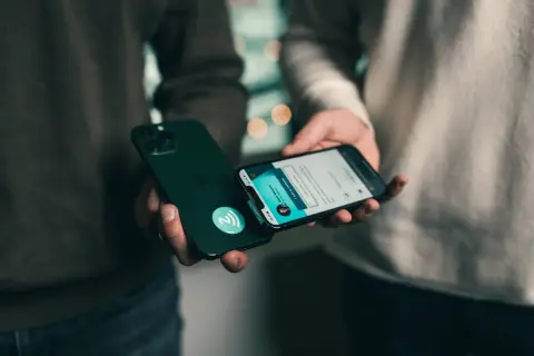 Dwie osoby zbliżają do siebie iPhone'y, jeden z nich ma naklejkę z symbolem NFC.