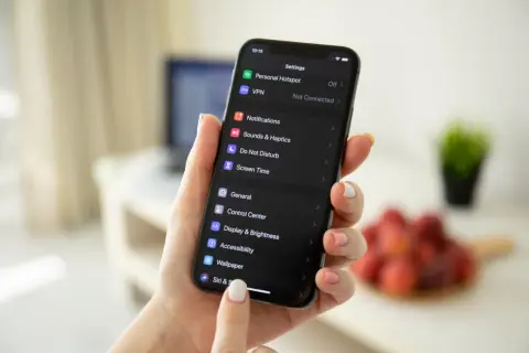 Dłoń trzyma iPhone'a z ciemnym motywem. Na ekranie widać ustawienia, w tym opcję "Wallpaper", która pomoże Ci dowiedzieć się, jak zmienić motyw na IG.