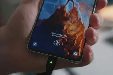 Dłoń trzyma smartfon z włączonym szybkim ładowaniem. Na ekranie widać informację o naładowaniu do 74% i szacowany czas do pełnego naładowania.