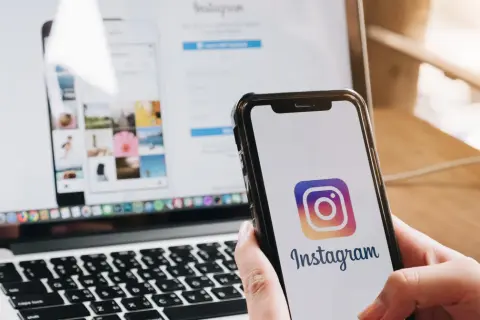 Dłoń trzyma smartfon z logo Instagrama. Obok laptop z otwartą stroną logowania, pokazującą jak wylogować się z Instagrama.