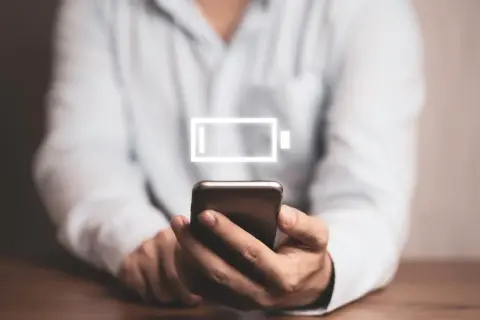 Osoba trzyma smartfon z ikoną niskiego poziomu baterii. Dowiedz się, jak wyłączyć optymalizację baterii Samsung, by dłużej cieszyć się energią.