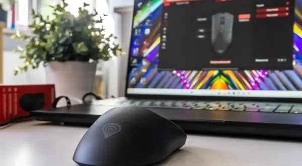 Czarna myszka komputerowa z logo Genesis na biurku, obok laptopa z kolorową tapetą i oprogramowaniem do konfiguracji. Wygląda jak sprzęt refurbished.