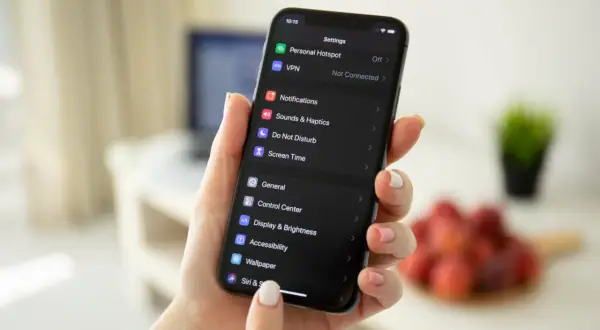 Dłoń trzyma iPhone'a z ciemnym motywem. Na ekranie widać ustawienia, w tym opcję "Wallpaper", która pomoże Ci dowiedzieć się, jak zmienić motyw na IG.