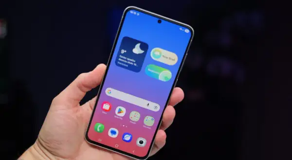 Dłoń trzyma smartfon Samsung z kolorowym ekranem. Widoczne widżety pogody i wiadomości, ikony aplikacji. Dbałość o ochronę baterii Samsung jest kluczowa.