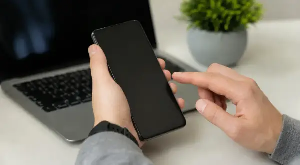 Dłoń trzyma smartfon, palec wskazujący dotyka ekranu, obok laptop. Może to być moment przed przywróceniem ustawień fabrycznych.