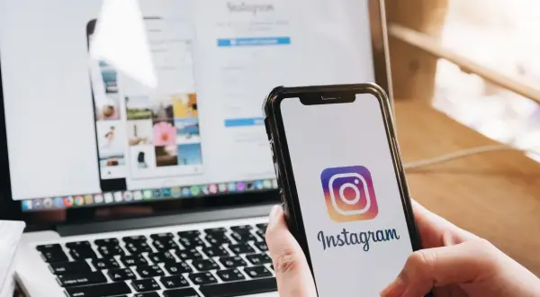 Dłoń trzyma smartfon z logo Instagrama. Obok laptop z otwartą stroną logowania, pokazującą jak wylogować się z Instagrama.