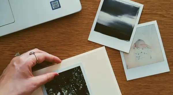 Dłoń z pierścionkiem układa zdjęcia na albumie. Obok leży laptop i kolejne fotografie, tworząc przestrzeń do przechowywania zdjęć.