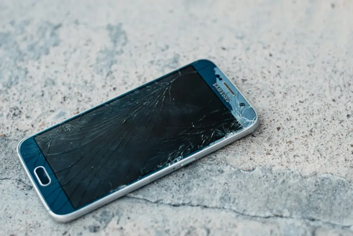 Rozbity ekran telefonu Samsung z widoczną pękniętą kreską na wyświetlaczu, leżący na betonowej powierzchni.