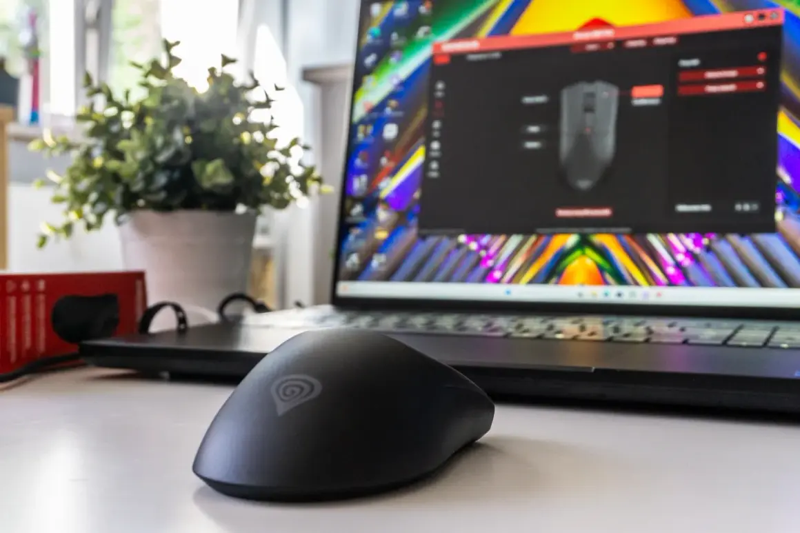 Czarna myszka komputerowa z logo Genesis na biurku, obok laptopa z kolorową tapetą i oprogramowaniem do konfiguracji. Wygląda jak sprzęt refurbished.