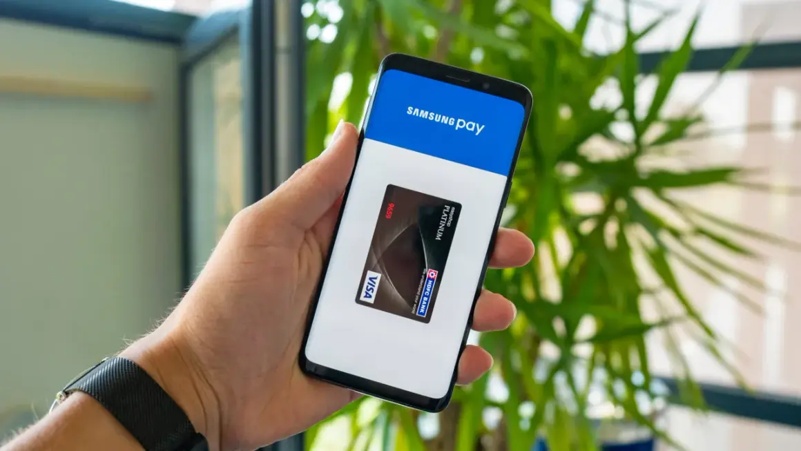 Dłoń trzyma smartfon z otwartą aplikacją Samsung Wallet, na ekranie widoczna karta płatnicza Visa.