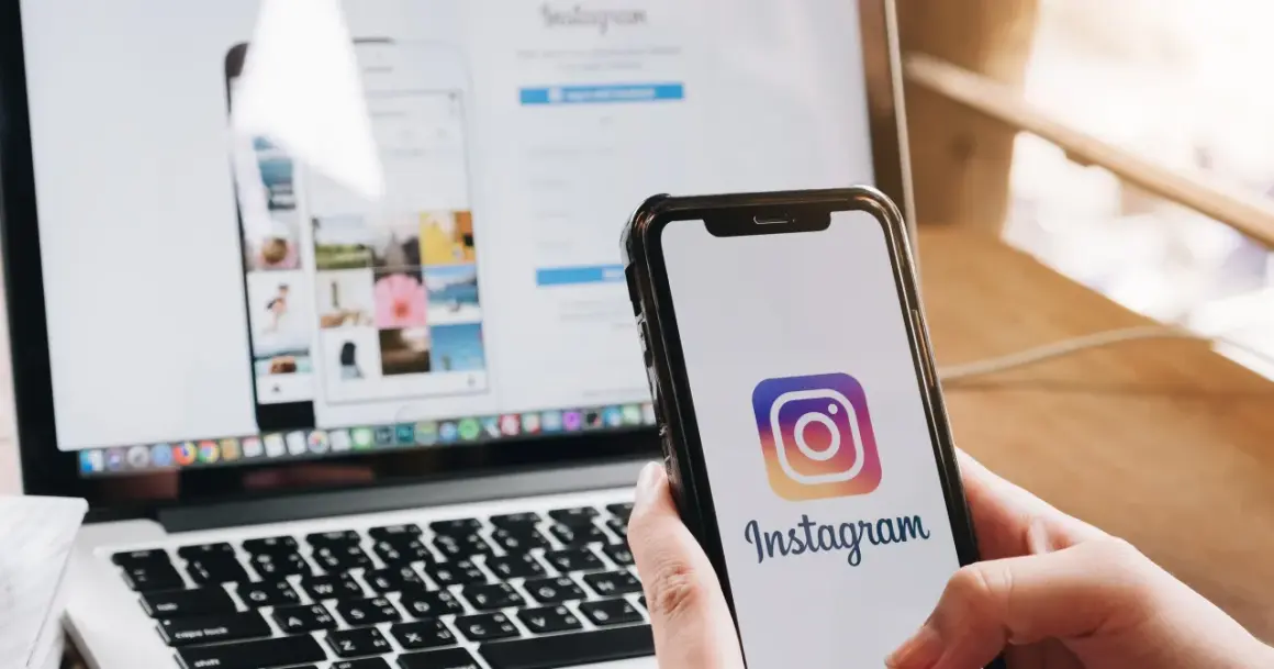 Dłoń trzyma smartfon z logo Instagrama. Obok laptop z otwartą stroną logowania, pokazującą jak wylogować się z Instagrama.