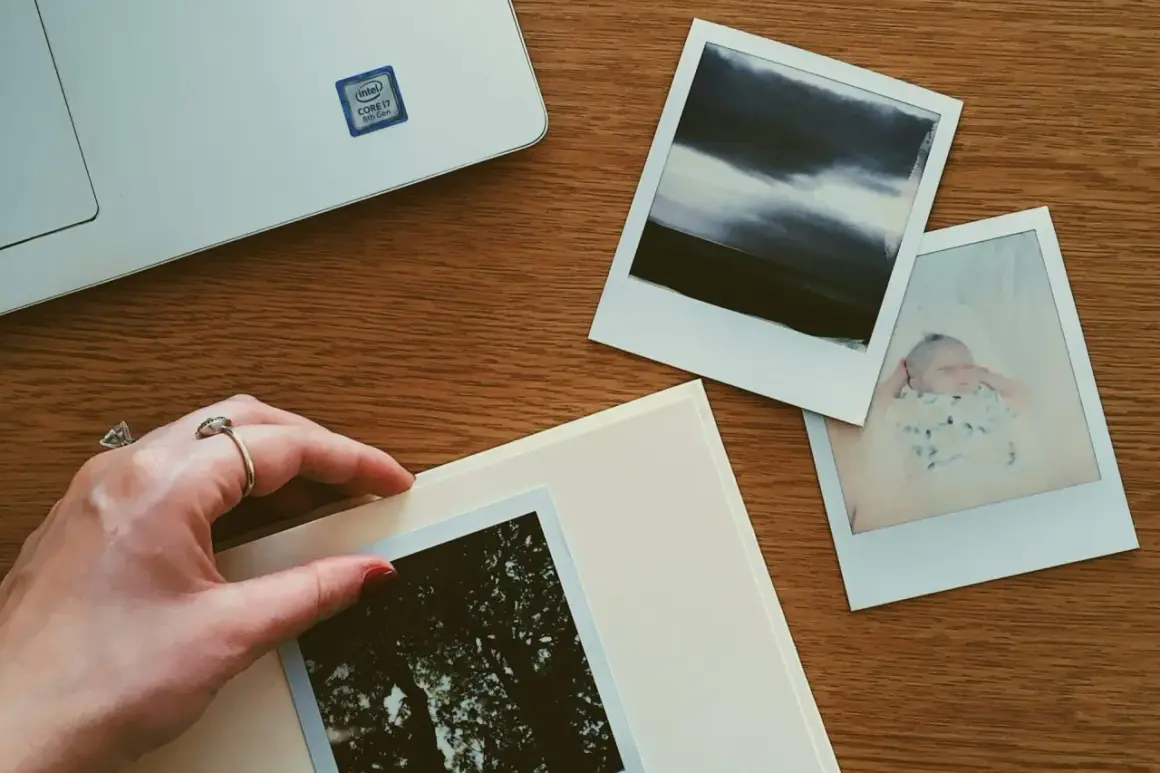 Dłoń z pierścionkiem układa zdjęcia na albumie. Obok leży laptop i kolejne fotografie, tworząc przestrzeń do przechowywania zdjęć.