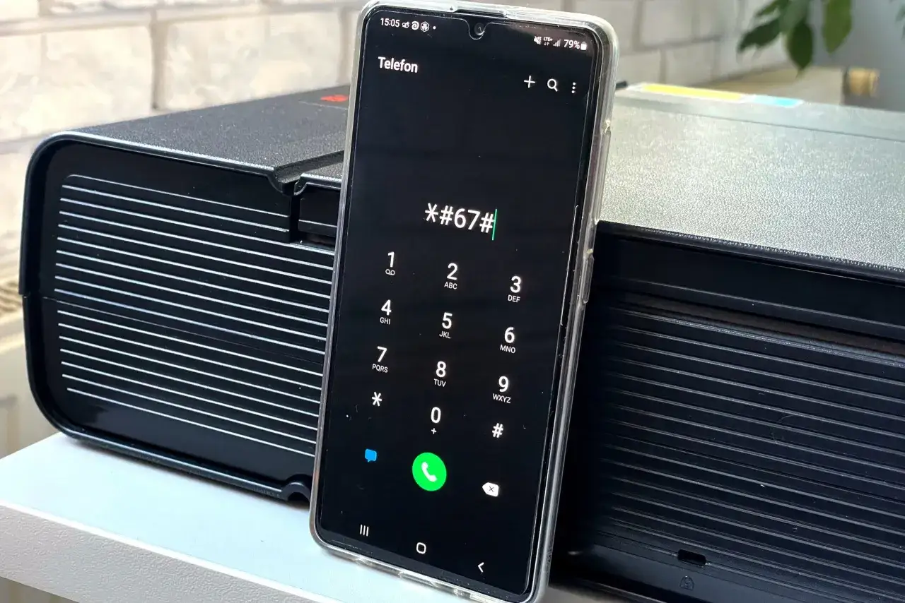 Telefon Samsung z wpisanym kodem *#67#. Urządzenie stoi na tle czarnych urządzeń.