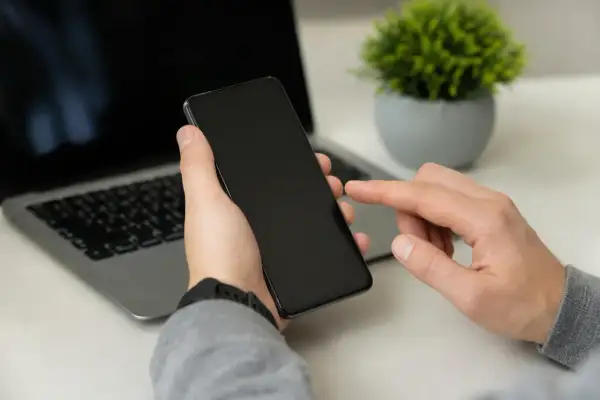 Dłoń trzyma smartfon, palec wskazujący dotyka ekranu, obok laptop. Może to być moment przed przywróceniem ustawień fabrycznych.